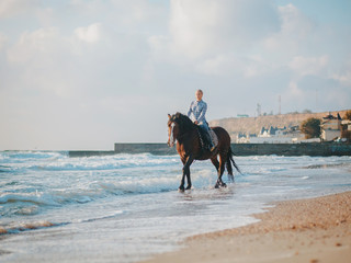 blonde woman sitting on black horse at sea beach. Beautiful woman wearing casual clothing enjoying sunrise morning close to ocean water.