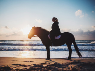 shof of woman sitting on black horse at sea beach. Beautiful woman wearing black clothing enjoying sunrise morning close to ocean water.
