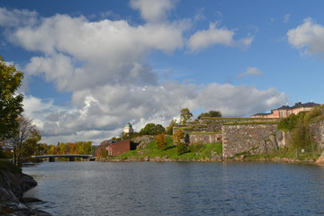 Stone Wall by Water with Blue Sky, Bridge,  Fluffy Clouds and Fall Color in Background
