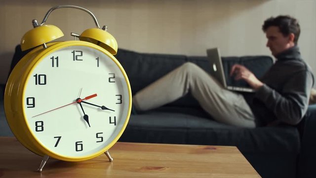 Young man using laptop sitting on couch at home. Big yellow clock is ticking