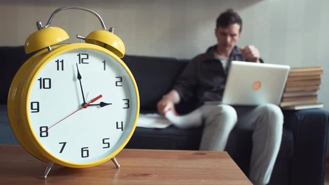 Young student using laptop sitting on couch at home. Big yellow clock is ticking