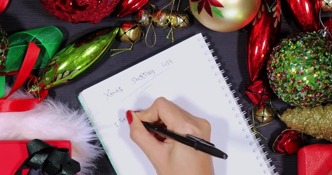 Top view of anonymous woman hand writing a shopping list foods and drinks for Xmas on notepad between Christmas ornaments on the table. Shot in 4k resolution