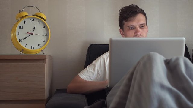 Young student using laptop sitting on couch at home. Big yellow clock is ticking