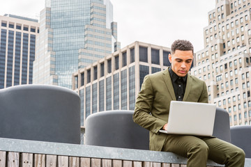 Young handsome American Businessman traveling, working in New York, wearing green suit, sitting at park in business district with high buildings, looking down, reading, working on laptop computer.