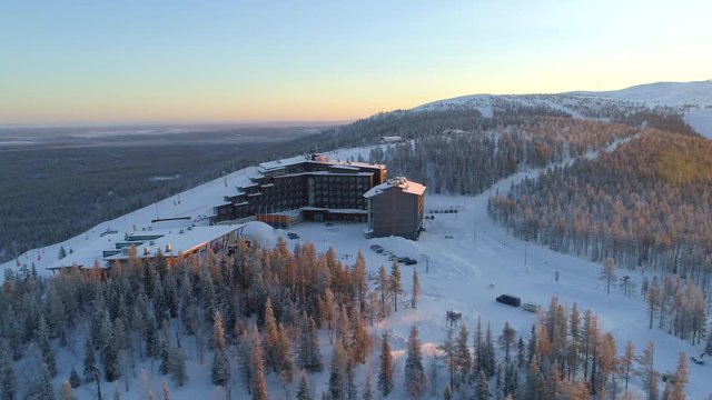AERIAL: Flying around hilltop skiing resort surrounded by ski slopes in winter. Snowy hotel on top of ski tracks at golden winter sunset. Winter sun rising above quiet ski resort Levi in Finland