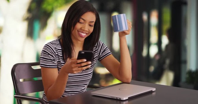 Cute black female sitting outdoors scrolls through phone laughing at pictures