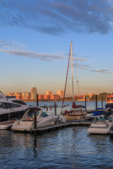 Power and sail boats docked on the Manhattan side of the Hudson River. The sun is rising and is hitting the buildings on the New Jersey side of the Hudson. There is a blue sky with pink clouds.