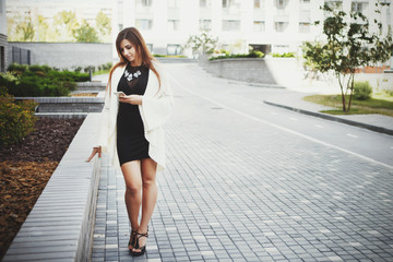 Beautiful young fashionable woman in a black short dress and white cardigan posing in the city. Fashion photo