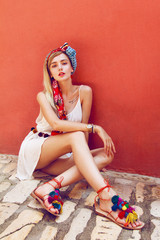 Beautiful woman sitting on a stone street leaning against a red wall. In her beautiful long straight blonde hair is styled colorful scarf. Wearing nice white dress and colorful sandals.