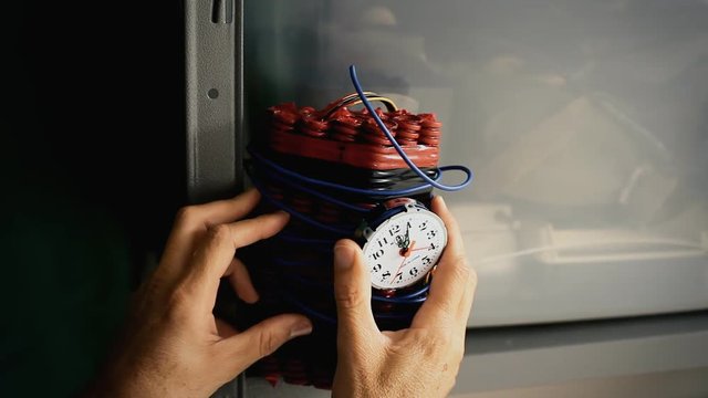 The hands of a man putting a cartoonish time bomb (TNT dynamite sticks with a timer clock) in a warehouse.
