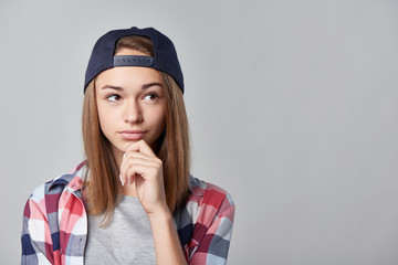 Closeup portrait of pensive teen girl wearing checkered shirt and baseball cap looking away at blank copy space, over grey background