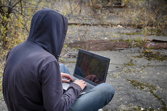 Hacker or programmer or cyber terrorist in hood sits with his back to us and working on laptop among the apocalyptic landscape of old destructed road with rusty construction in back of image. HDR