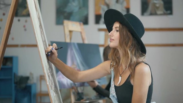 Smiling female artist, in a black stylish hat, drawing a picture with her colleague painting in the background, slow indoor motion