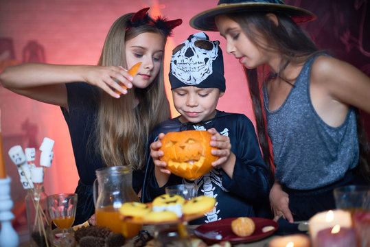 Portrait of three happy children wearing Halloween costumes, two girls and one boy holding carved pumpkin looking into it during party in decorated room
