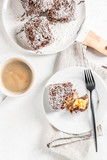 Australian food. Traditional dessert Lamington - pieces of biscuit in dark chocolate, sprinkled with coconut powder chips. On a marble plate, white table. With coffee mug. Copy space top view