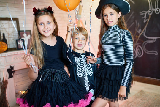 Portrait of three happy children wearing costumes posing with balloons in decorated room celebrating Halloween
