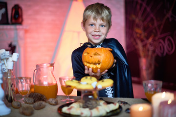 Waist-up portrait of handsome little boy wearing skeleton costume holding Halloween scary pumpkin in hands while posing for photography, interior of decorated room on background