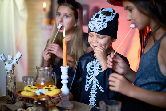 Portrait of handsome little boy wearing skeleton costume enjoying tasty fried marshmallows while celebrating Halloween with female friends, interior of decorated room on background