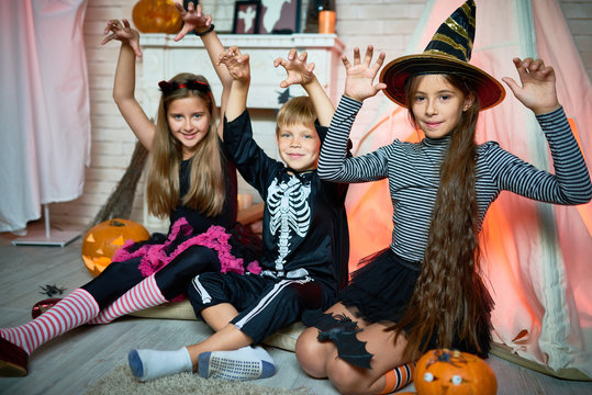 Group portrait of joyful children wearing Halloween costumes posing for photography while sitting on wooden floor of living room decorated for holiday