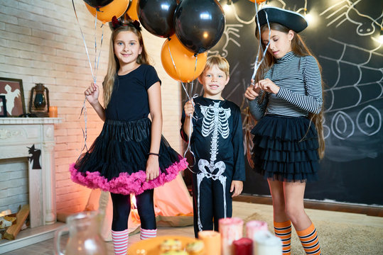 Group portrait of smiling little friends wearing fancy costumes holding bunches of balloons in hands while posing for photography, interior of living room decorated for Halloween on background