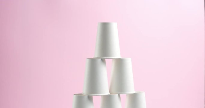 Young woman builds and breaks tower pyramid of white paper cups turned upside down on pink background