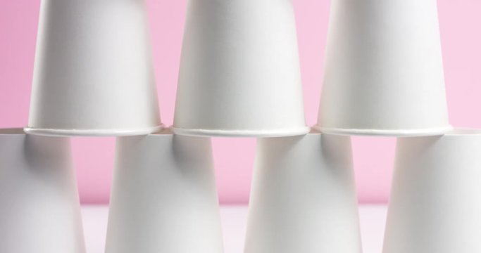 Young woman builds and breaks tower pyramid of white paper cups turned upside down on pink background