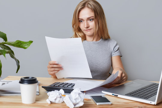 Horizontal portrait of successful businesswoman reads attentively business papers or contract, calculates everything, surrounded with modern gadget, drinks takeaway coffee. Career, business concept