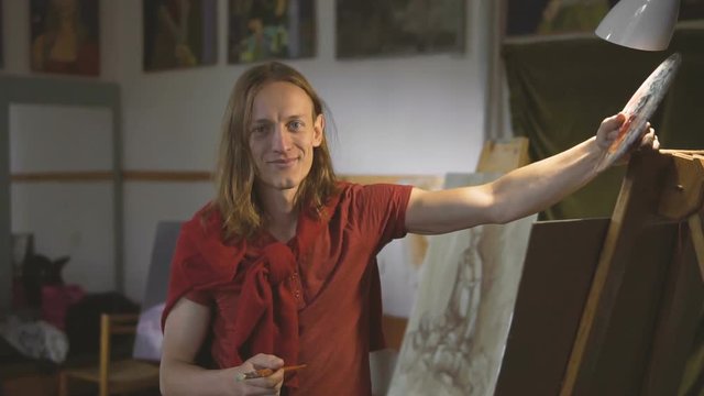 Long hair caucasian male painter smiles standing near the easel in his studio, slow indoor motion