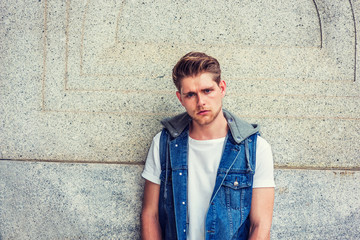 Unhappy Young American College Student with little beard in New York, wearing blue Denim hoody sleeveless vest jacket, white T shirt, standing against vintage wall on campus, frowned, sad, thinking. .
