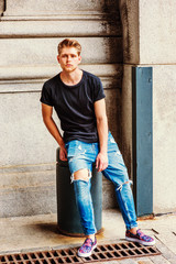 Young Modern American Man with little beard thinking outside in New York, wearing black T shirt, destroyed jeans, fashionable shoes, sitting on metal pillar on vintage street, relaxing..