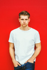 Portrait of Young Handsome Man with little beard in New York, wearing white T shirt, blue jeans, hands in pocket, standing against red background, seriously looking at you..