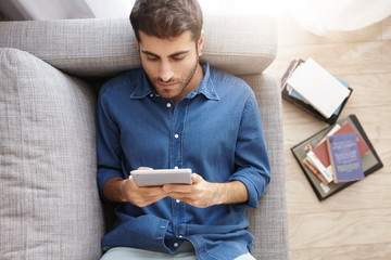 Relaxed unshaven male in formal shirt lies on comfortable couch, watches new film on touch pad using free internet connection, plays games online, enjoys conversation with friends in social networks © WHstudio Leushin N
