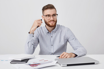 Fototapeta premium Portrait of male office worker, has specific appearance, dressed formally, sits at white desk, uses laptop, smart phone and documents for work, looks aside as notices his best companion or colleague