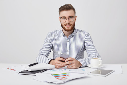 Isolated stdio shot of pleasant looking stylish male manager with stubble, holds smart phone as messages online, uses high speed internet connection, drinks coffee, rests after studying documents