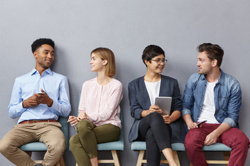 Horizontal portrait of people sit in queue, have pleasant conversation with each other, share ideas and life experience, isolated over grey concrete wall. Diverse group in row, speak and hold gadgets - Powered by Adobe