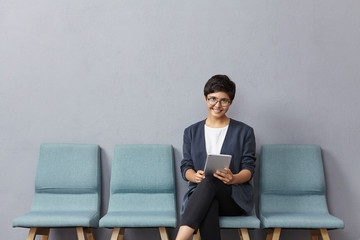 Pleasant looking mixed race woman has short trendy hairstyle, wears spectacles and formal jacket, comes to job interview, waits in corridor, uses modern tablet computer for reading tips to succeed