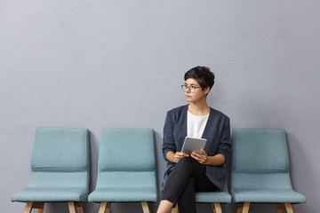 Cute businesswoman looks pensively aside, waits meeting with partners, sits on chairs, holds tablet for communication or creating project. Female candidate on manager position waits for interview