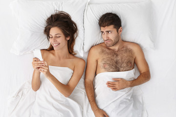 Obraz na płótnie Canvas Unhappy young bearded man feeling sad disappointed because of girlfriend doesn't pay attention to him. Attractive happy female enjoying online life, ignoring husband who is lying in bed next to her