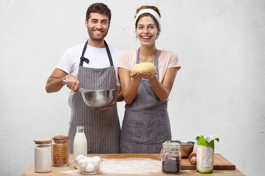 Products, Food, Cuisine And Cooking Concept. Portrait Of Happy Positive Young European Couple Baking Homemade Bread: Bearded Guy Beating Eggs In Bowl While Cute Woman Holding Round Shaped Dough