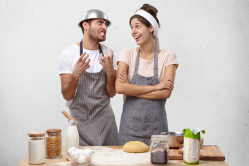 Young couple having quarrel while cooking in kitchen. Angry beadred guy with pan on head having irritated look, telling his wife to stop annoying him, pretty woman stnding next to him and smiling