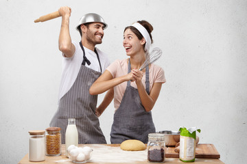 Happy female and male foolish at kitchen, fight with whisk and rolling pin, have glad expressions. Overjoyed cooks make fools, rest after making dough or pastry, stand near table with products