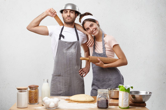 Smiling female model in apron leans at husband`s shoulder, recieves help and support from him. Happy successful glad woman and serious man make dough, use rolling pins, bake pastry or delicious pie