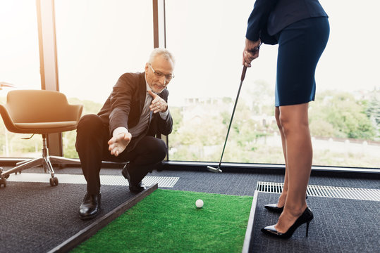 A Woman In A Black Business Suit Plays Golf In The Office. An Old Man In A Business Suit Helps Her