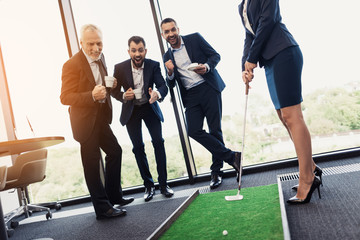 Three businessmen and business ladies play golf. A woman is holding a golf stick in the dots. Men are watching