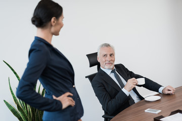The girl secretary is flirting with her boss. He sits at his desk with a cup of coffee in his hands