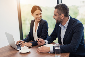 A man and a woman in strict business suits are discussing something while sitting at a table with a cup of coffee