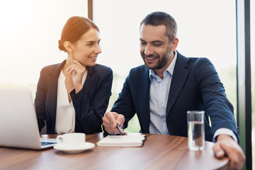 A man and a woman in strict business suits are discussing something while sitting at a table with a cup of coffee