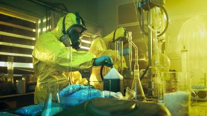 In the Underground Laboratory Two Clandestine Chemists Wearing Protective Coveralls and Masks Cook Drugs. They Work with Beakers, Distillation Glassware, Canisters and Hosepipe. True Crime Concept.