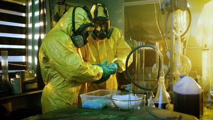 In the Underground Laboratory Two Clandestine Chemists Wearing Protective Masks and Coveralls Pack...