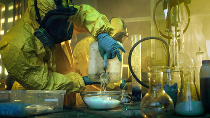 In the Underground Drug Laboratory Two Clandestine Chemists Covered in Protective Coveralls and Gas...